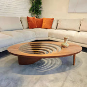 Modern oval glass and wood coffee table with sculptural base and layered glass inlay, styled in a cozy living room with a light sectional sofa and neutral décor.